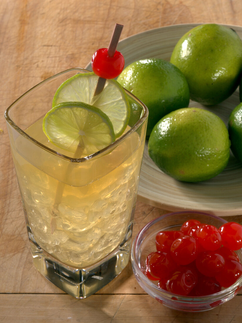 Close-up of cocktail with fruit skewer and bowls of cherries besides limes