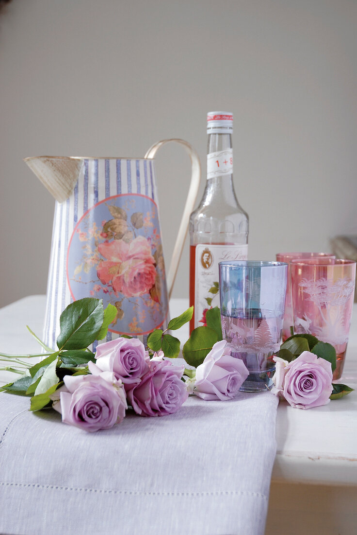 Jug with printed rose, bottle, coloured glasses and purple roses on white cloth