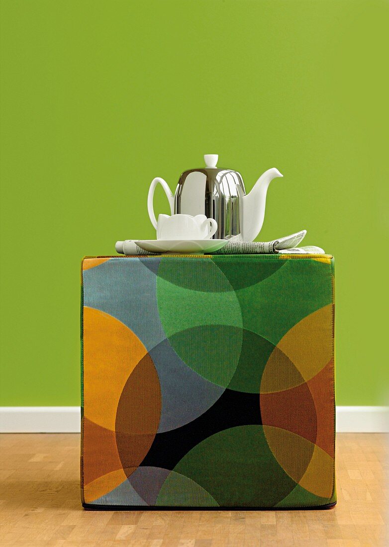 Teapot and teacup on multicoloured, retro-style pouffe against green wall
