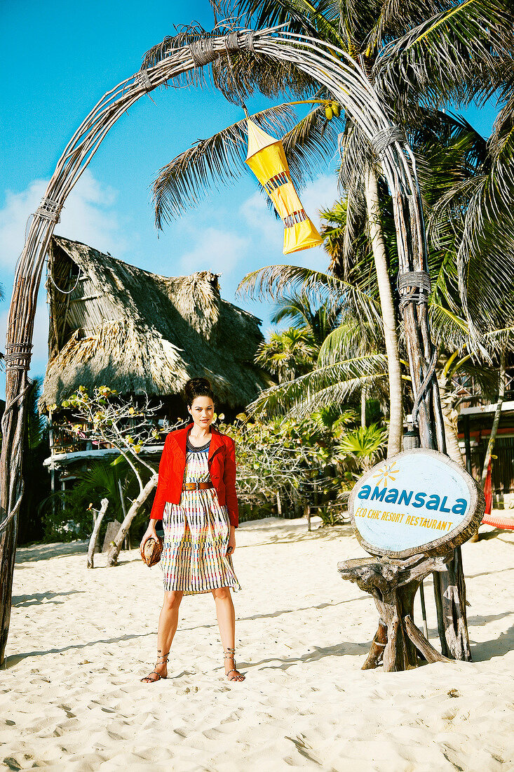 Pretty woman wearing jacket and colourful stripe dress holding coconut on beach