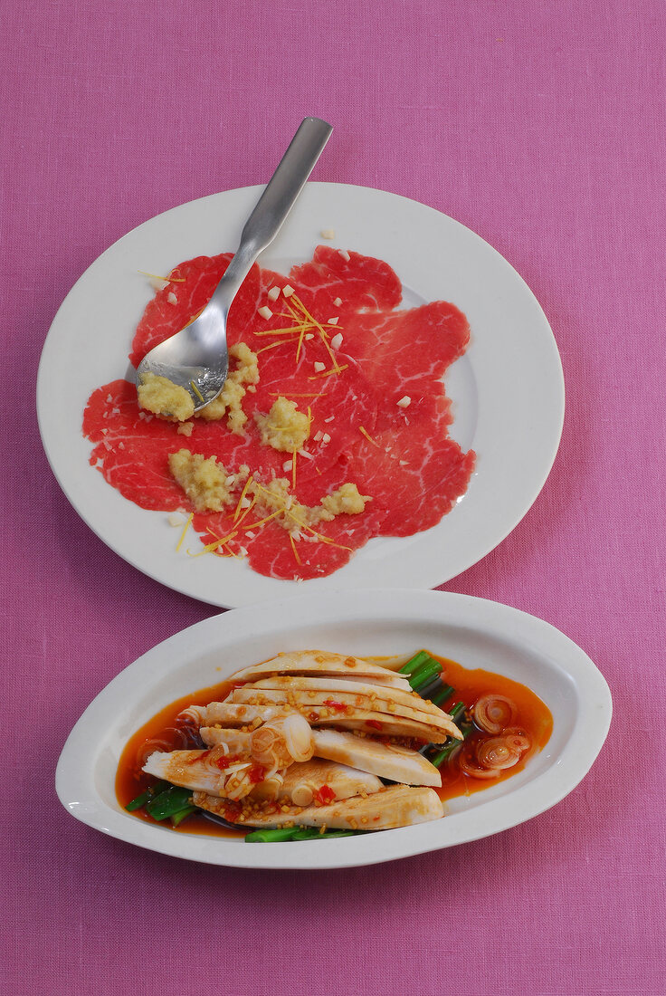 Carpaccio on plate and spicy chicken in serving dish on pink background