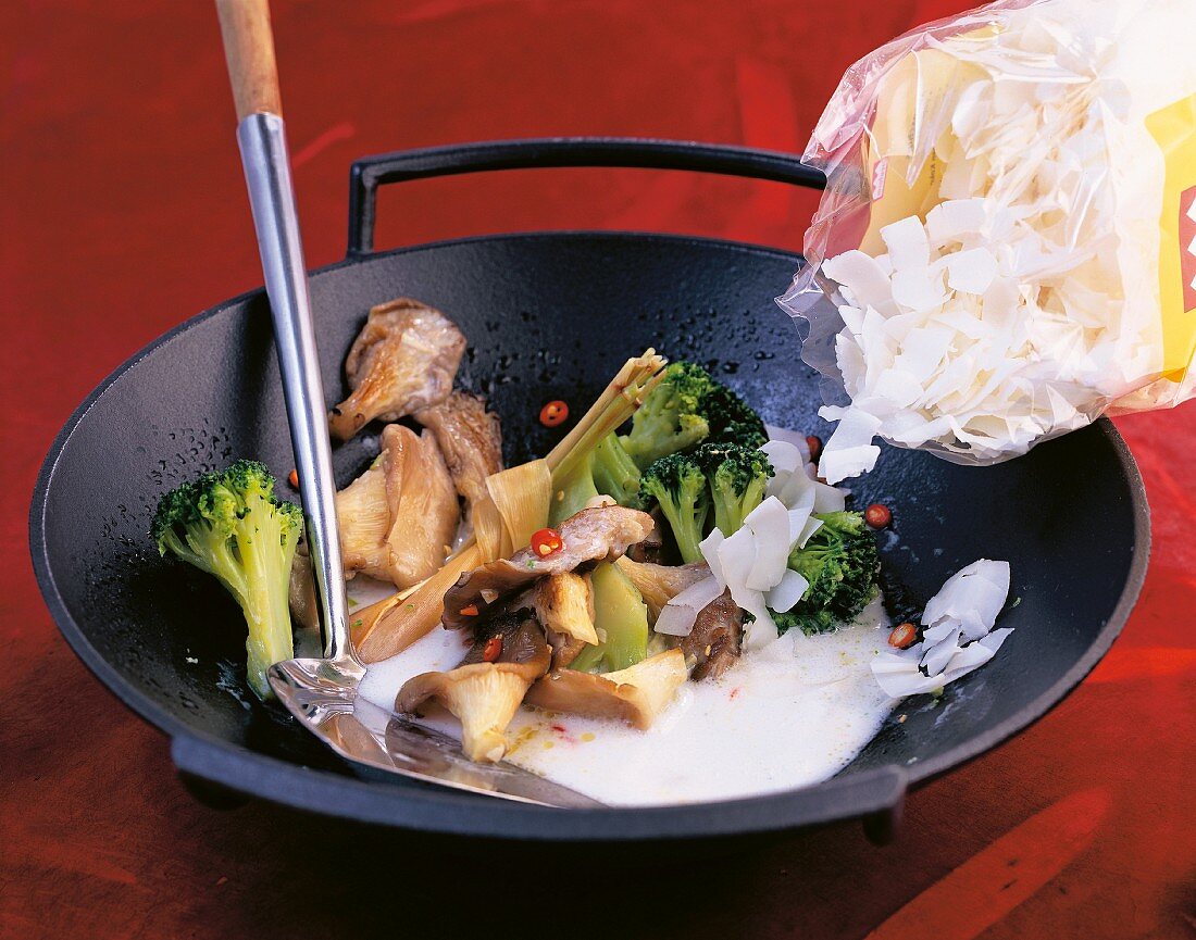 Coconut chips being added to broccoli, mushrooms and coconut milk in a wok