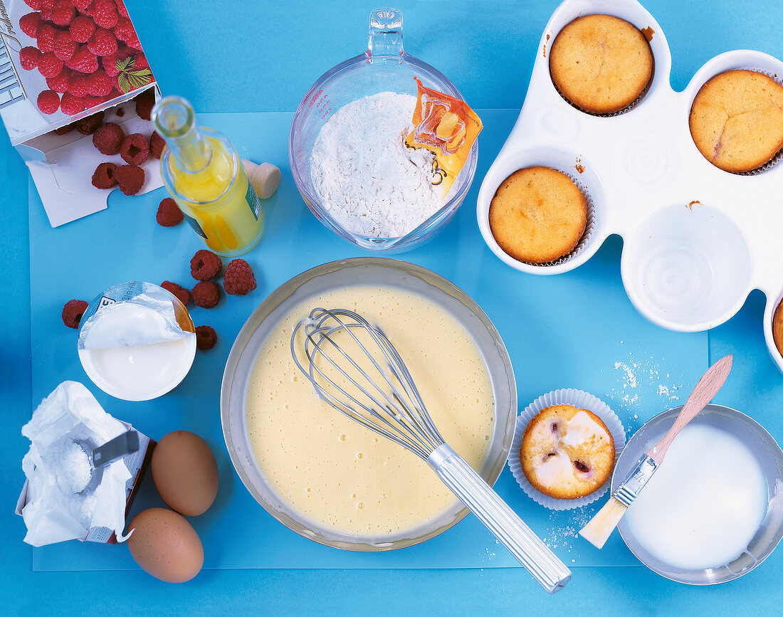 Batter and other ingredients for preparation of muffins on blue surface, overhead view