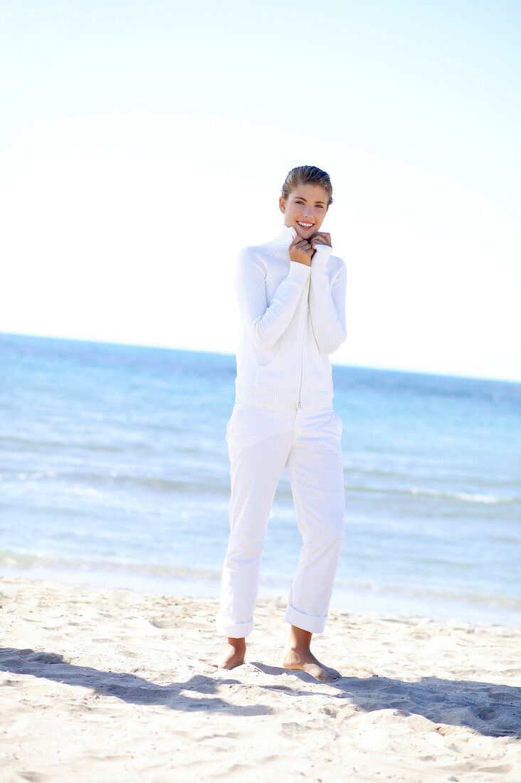 Woman in white outfit holding collar of the top while standing bare foot on beach, smiling