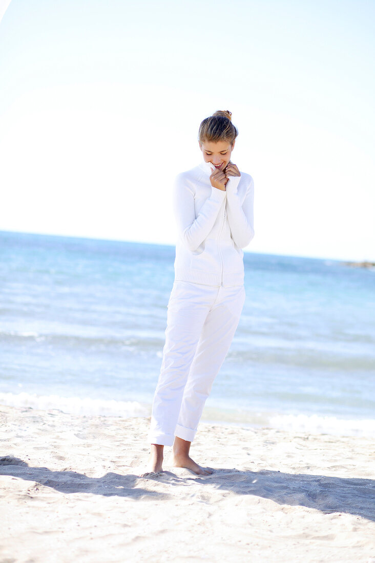 Shy blonde woman in white outfit standing bare foot on beach, looking down and smiling
