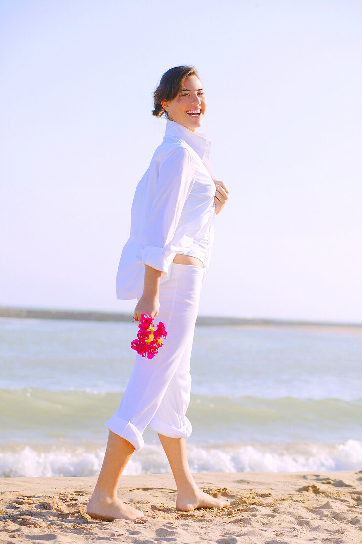 Cheerful brunette woman wearing white outfit walking on beach with flowers, smiling
