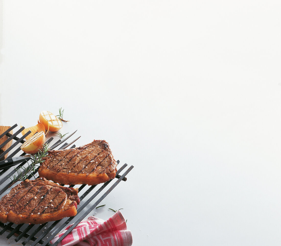 Steaks on grill rack with onions and herbs on white background, copy space