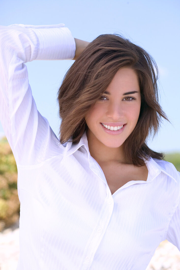 Portrait of pretty brunette woman wearing white blouse with hand behind her head, smiling