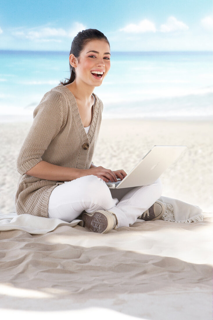 Portrait of beautiful woman sitting on sand and working on laptop, smiling widely