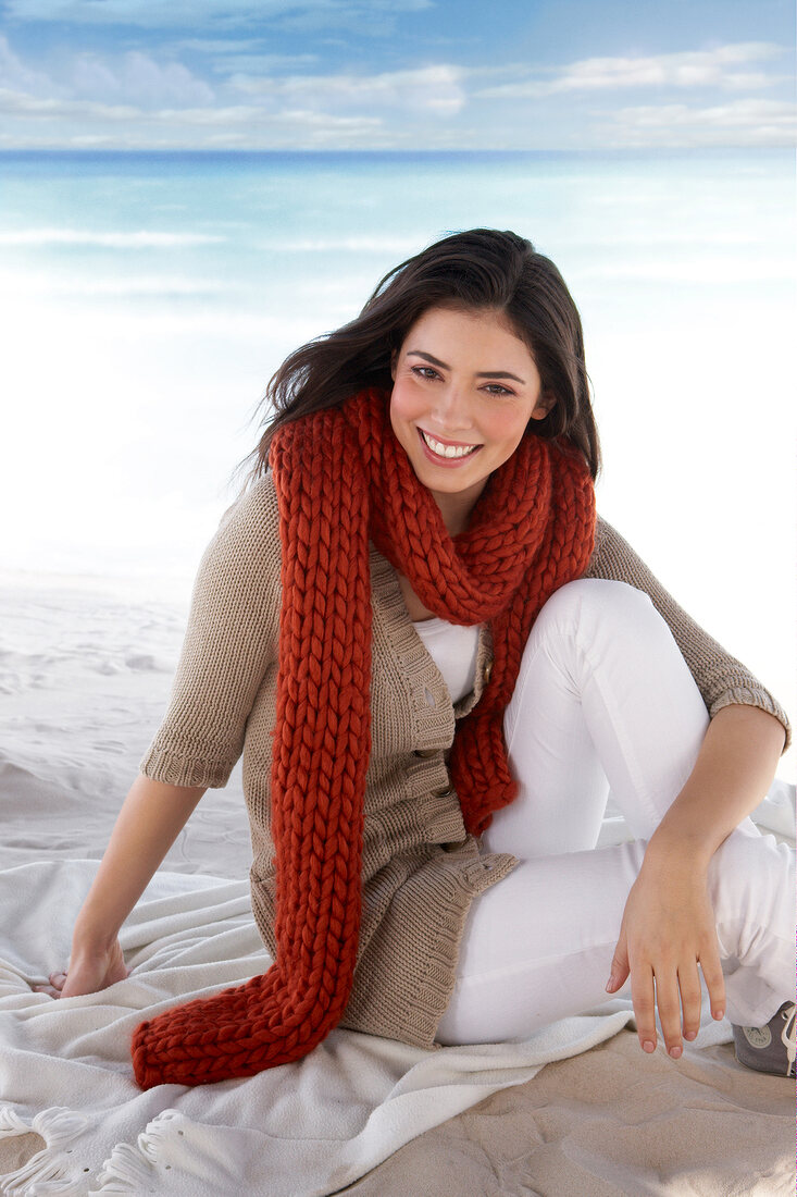 Portrait of beautiful woman wearing beige sweater and red scarf sitting on beach, smiling