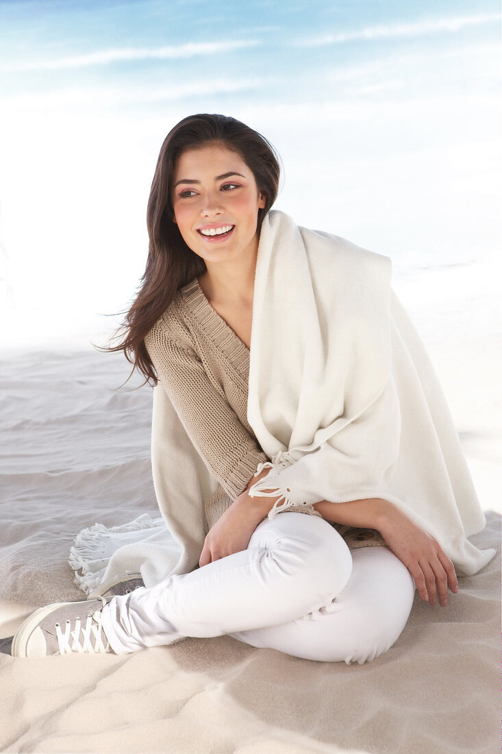 Beautiful woman wearing beige cardigan and white blanket sitting on beach, smiling