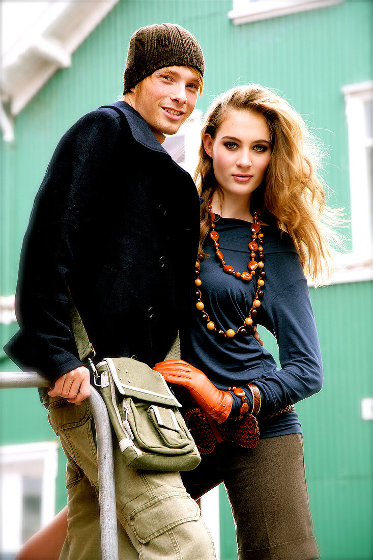 A couple in autumnal fashion standing on a railing in front of a green house