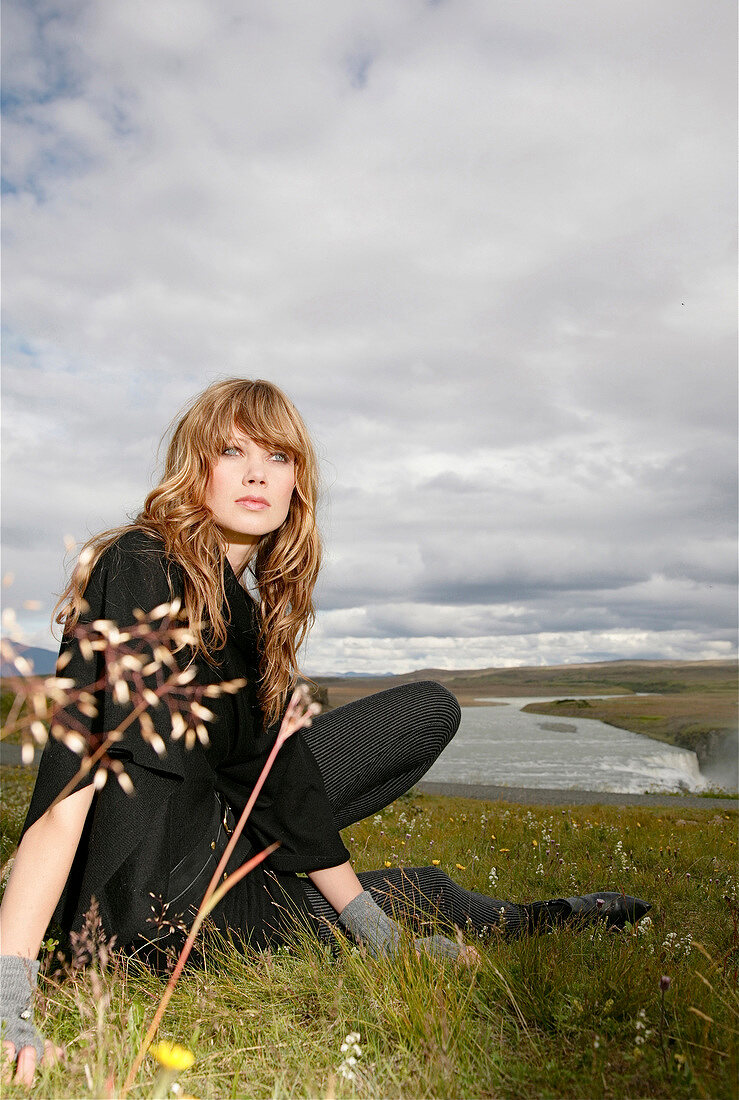 Pensive blonde woman with bangs wearing black top sitting on grass and looking away