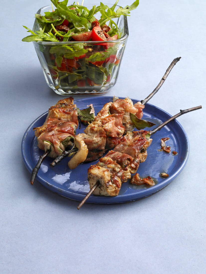 Saltimbocca skewers on plate with tomato salad in glass bowl
