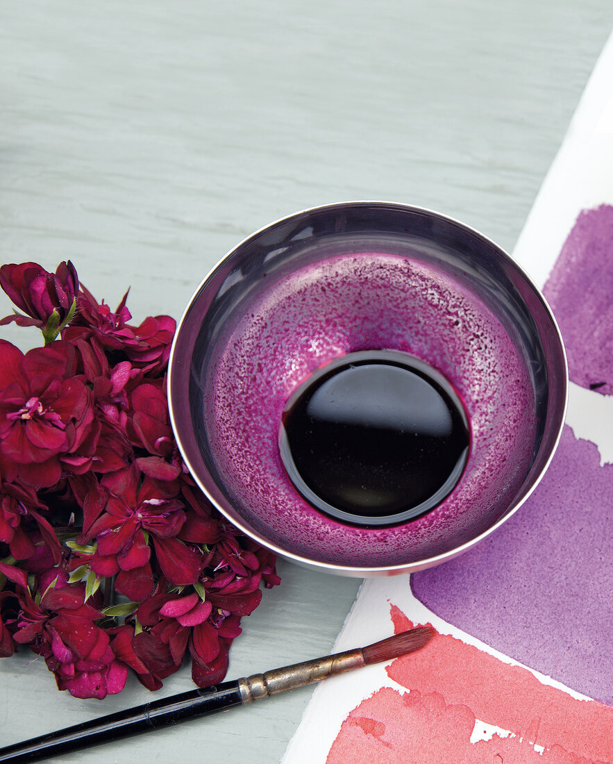 Purple colour with water in bowl with paint brush and geraniums flowers by side