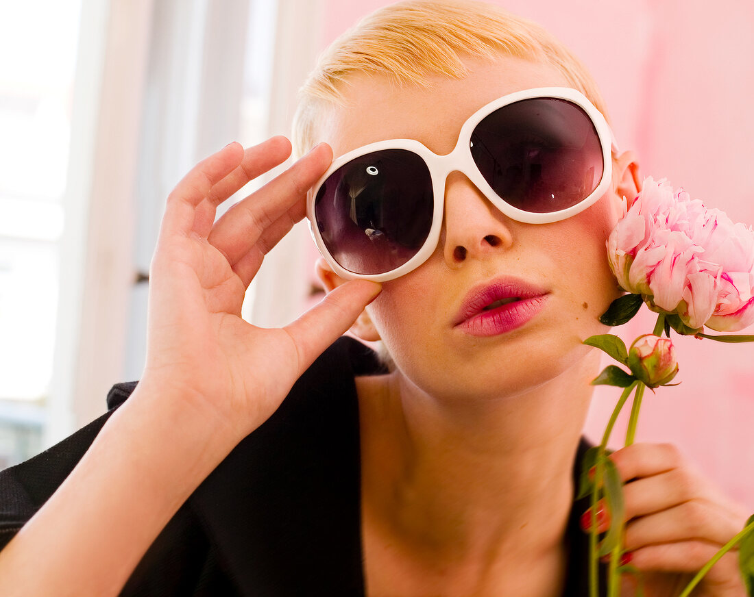 Close-up of blonde woman with short hair wearing white frame sunglasses holding flower