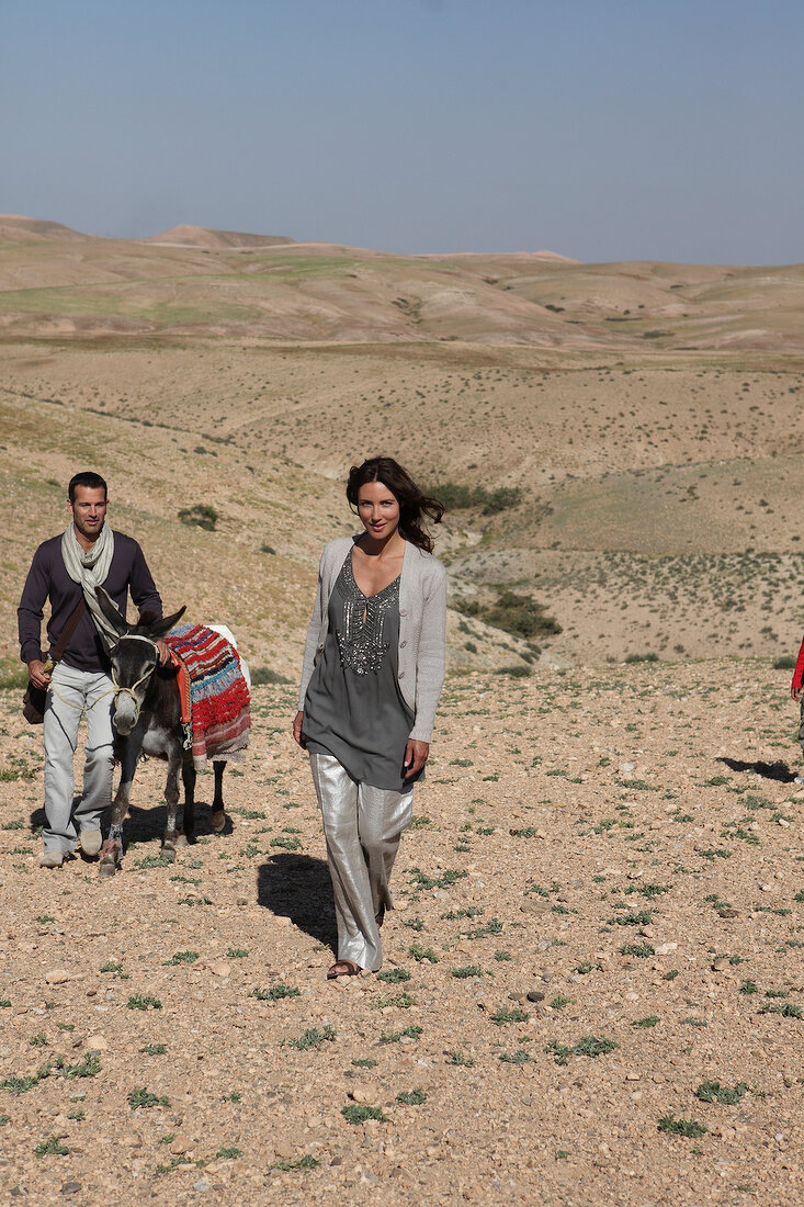 Woman wearing gray sweater and pants walking with man with donkey in desert