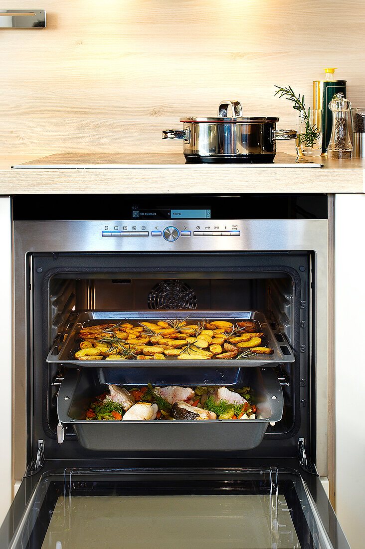 Baking tray with potatoes and fish in the oven with door open