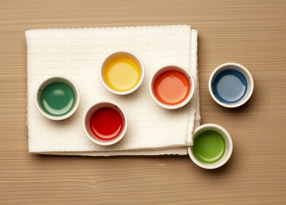 Bowls of various liquid paint and white cloth on wooden surface, overhead view