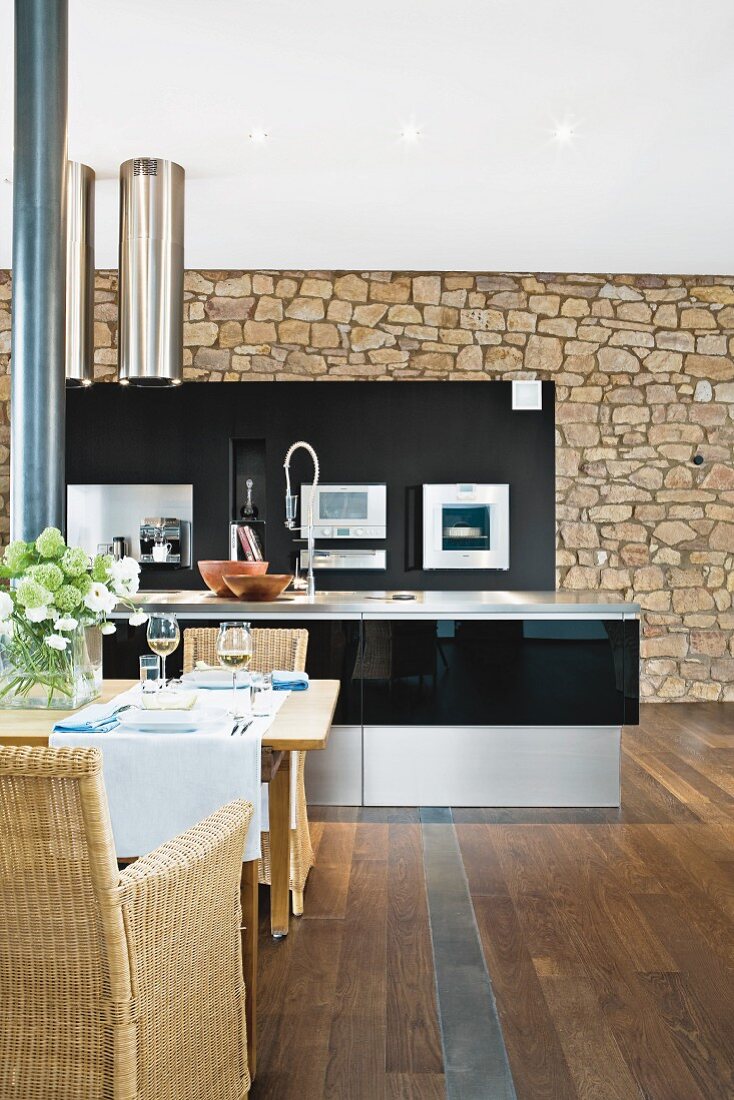 Black kitchen in front of stone wall; dining area with wicker chairs in foreground