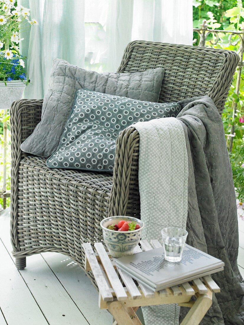 A grey wicker armchair with decorative cushions and blankets in various shades of grey on a summery balcony