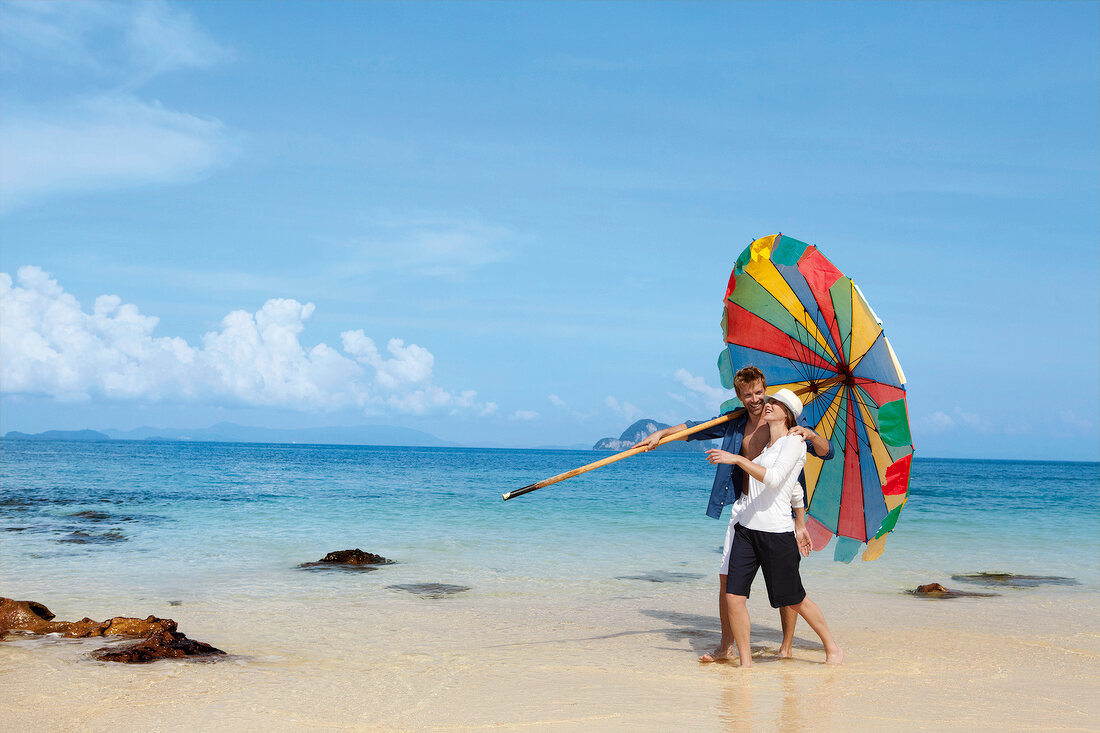 Couple holding multi coloured umbrella and walking on beach