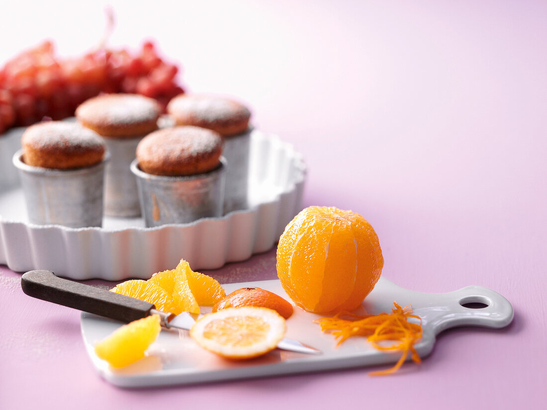 Eine Orange wird geschält, Cupcakes auf einem Tablett im Hintergrund