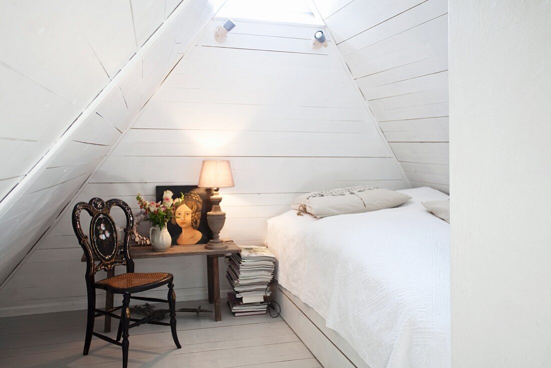 A bedroom in white in an attic with a bed, an antique wooden chair and a bedside table