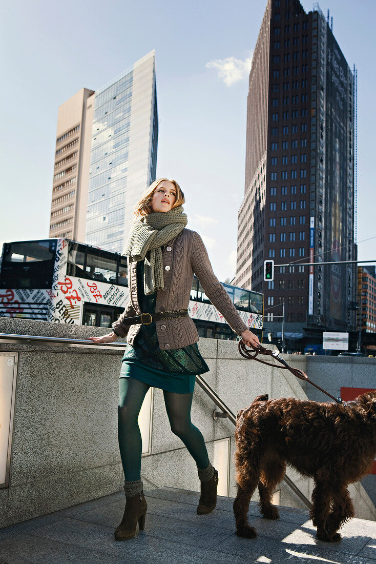 Fashionable woman wearing sweater over dress holding dog with a leash near staircase