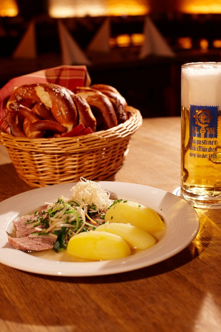 Meat, potatoes and radish on plate and basket of pretzels on wooden table