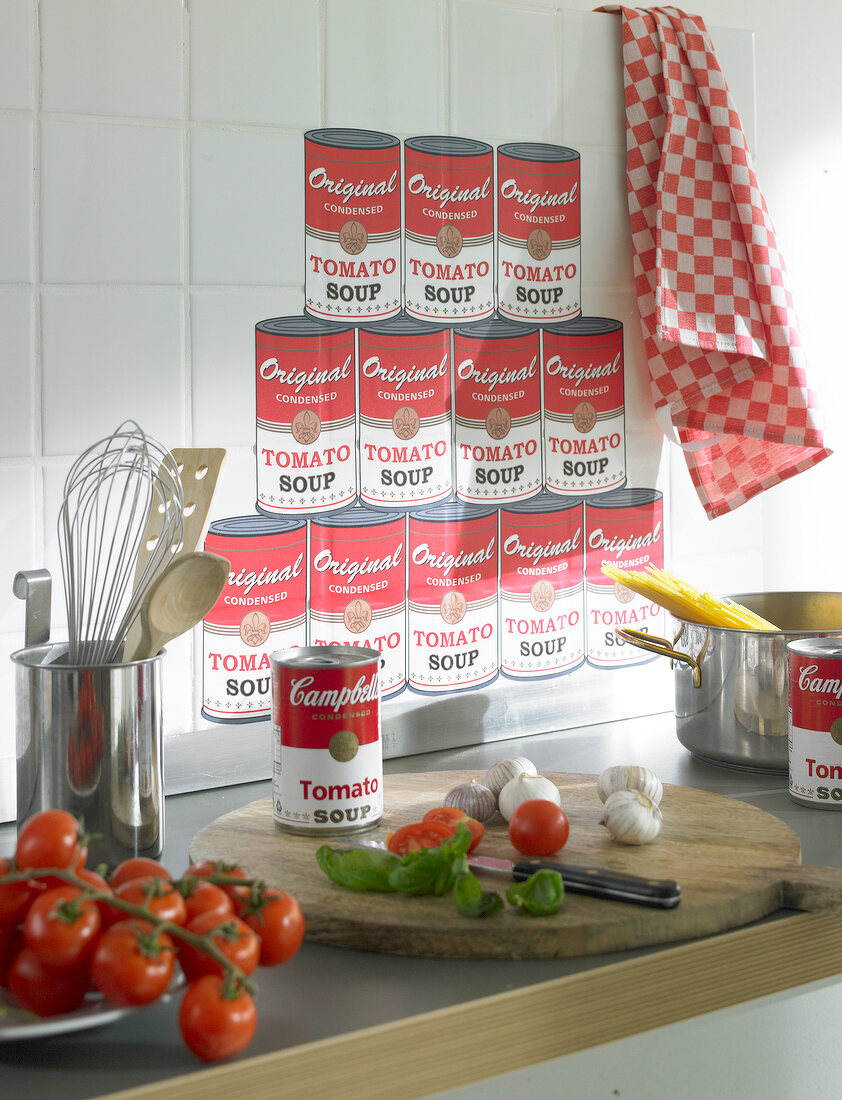 Chopping board with tin, vegetable and knife against tomato soup stickers on wall