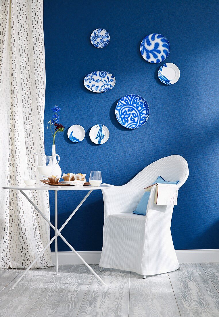 Kleiner Essplatz mit Klapptisch und Hussensessel vor Wand mit blauer Tapete und blau-weissen Wandtellern