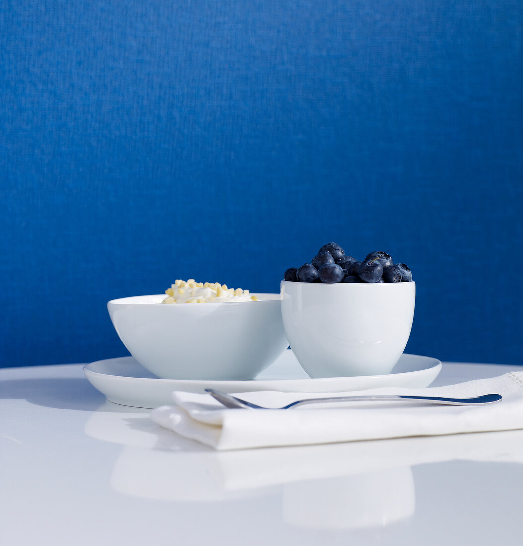 Two white bowls of fruits on table against blue wallpaper