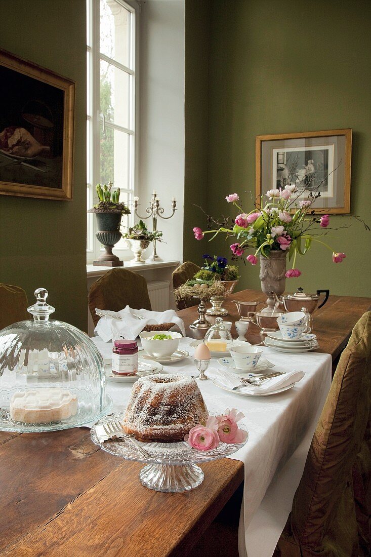 A long breakfast table with cake on a cake stand and under a glass cloche in a a traditional dining room with green-painted walls
