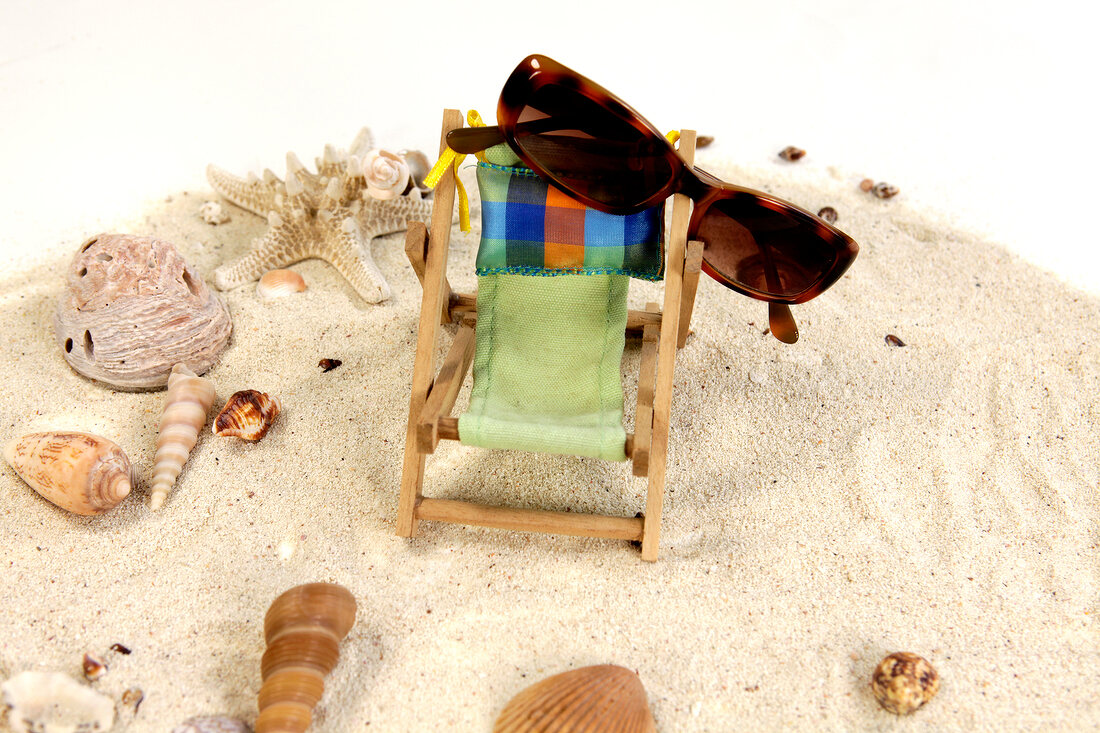 Close-up of sunglasses on deck chair surrounded by sea shells on sand