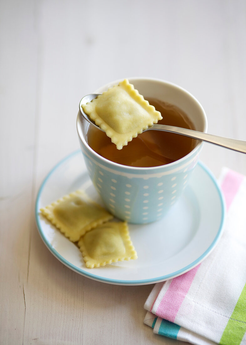 Cup of beef broth with ravioli on spoon and saucer