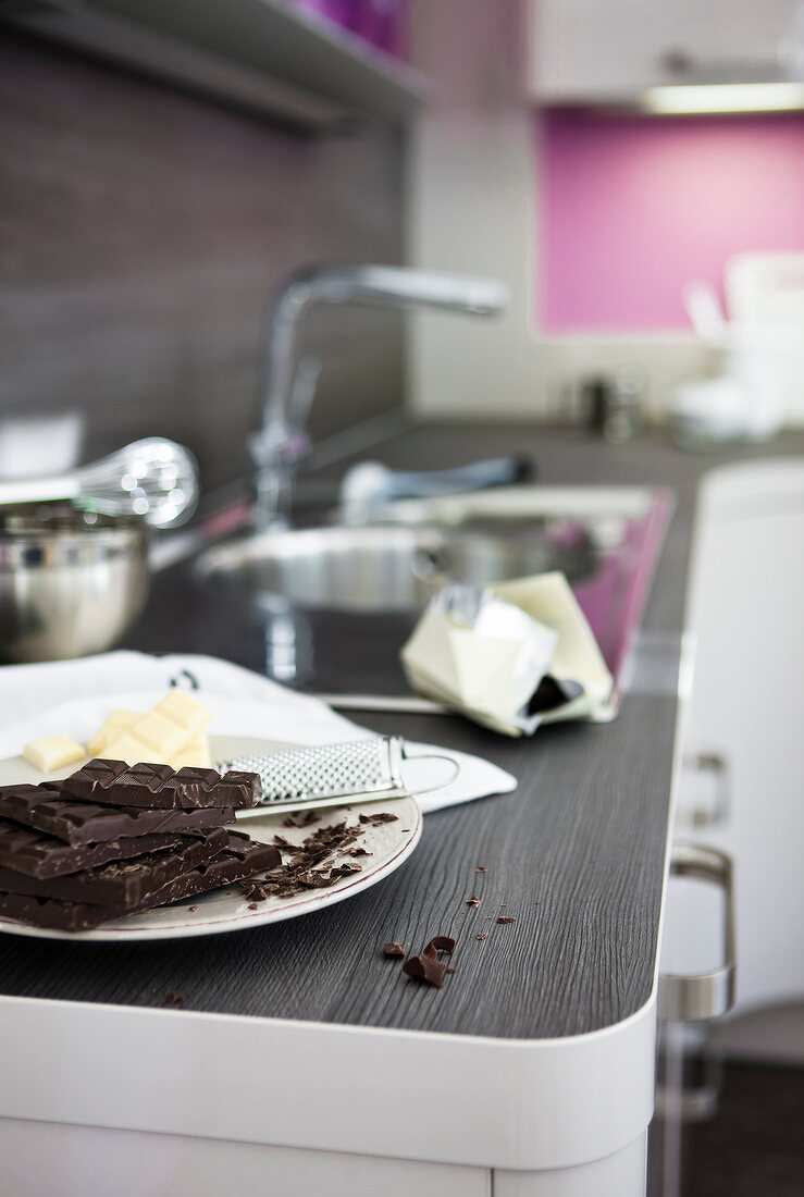Schokoladenstücke, auf Teller, mit Küchen-Raspel, auf Arbeitsplatte