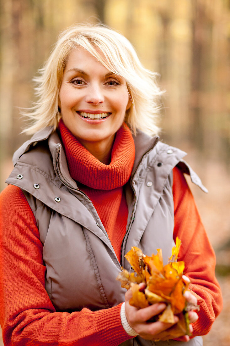 Happy blonde woman wearing gray jacket over turtleneck sweater, holding leaves and smiling