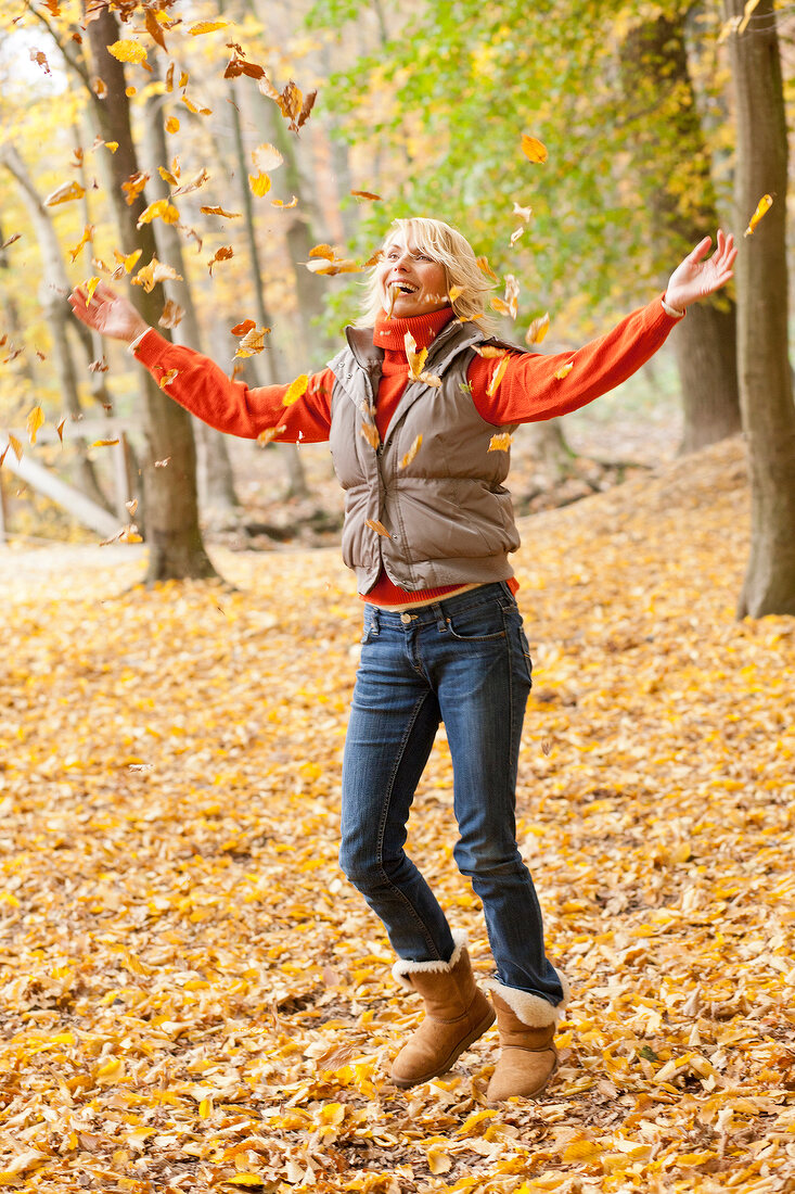 Excited blonde woman wearing gray jacket and jeans playing with autumn leaves in forest