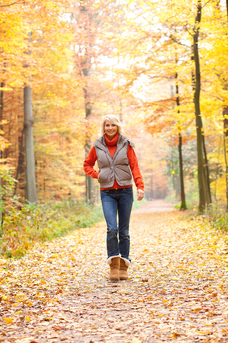 Blonde woman in gray jacket and jeans walking on forest path covered with autumn leaves