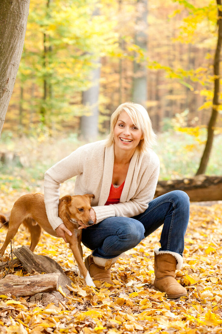 Pretty blonde woman in beige cardigan crouching and playing with dogs in forest, laughing
