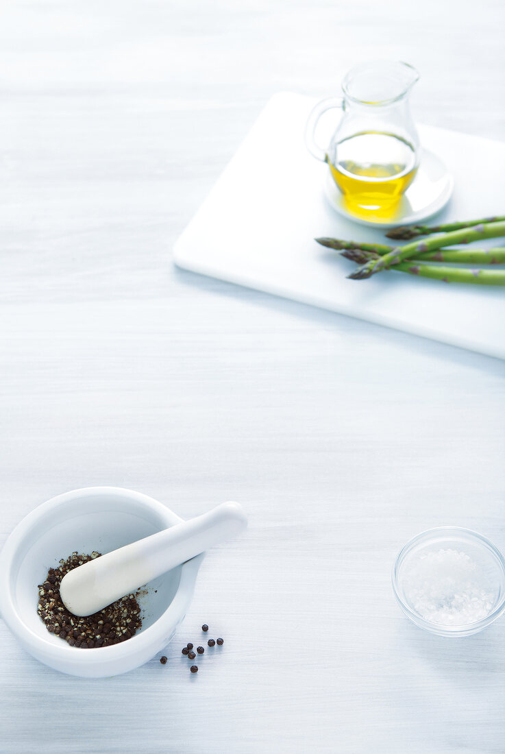 Spice in mortar, bowl with salt, oil and asparagus on white chopping board