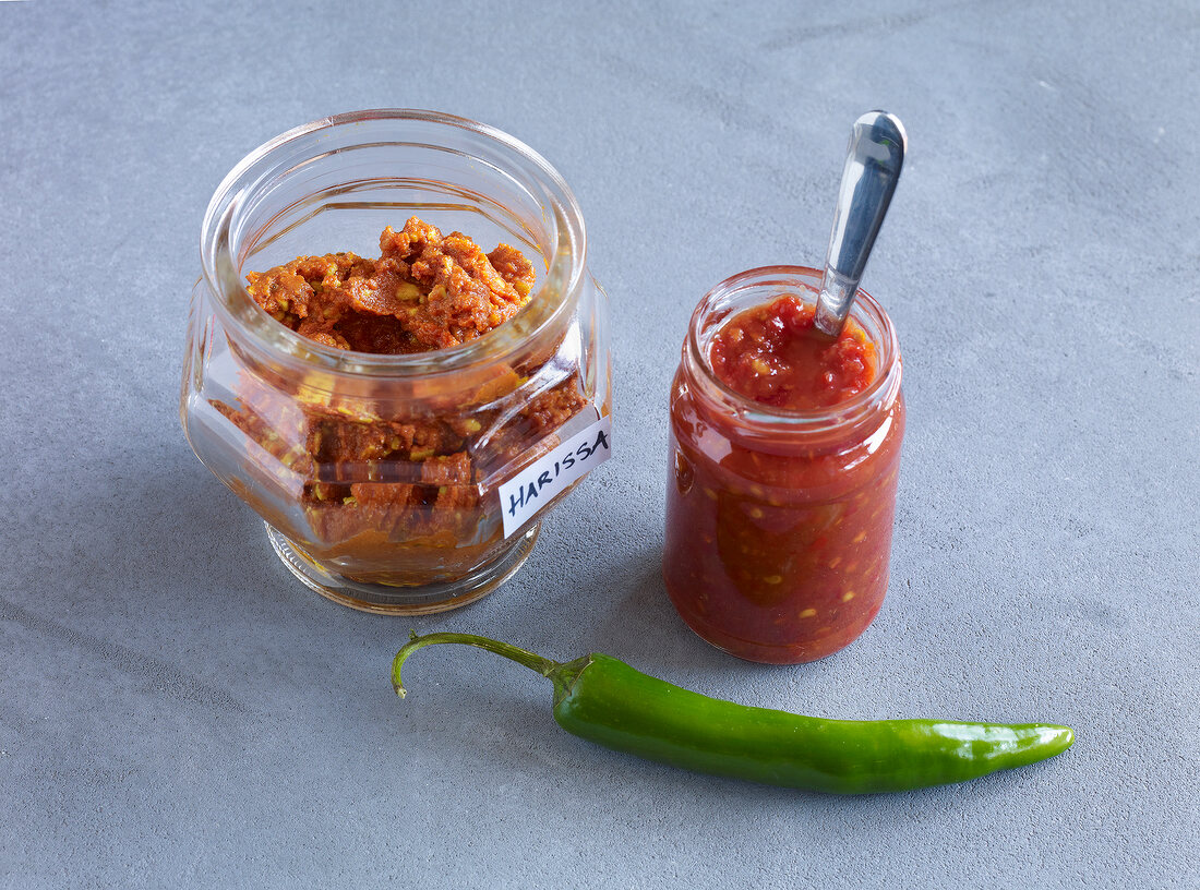 Harissa and sambal oelek in glass jars with green chilli pepper on gray background