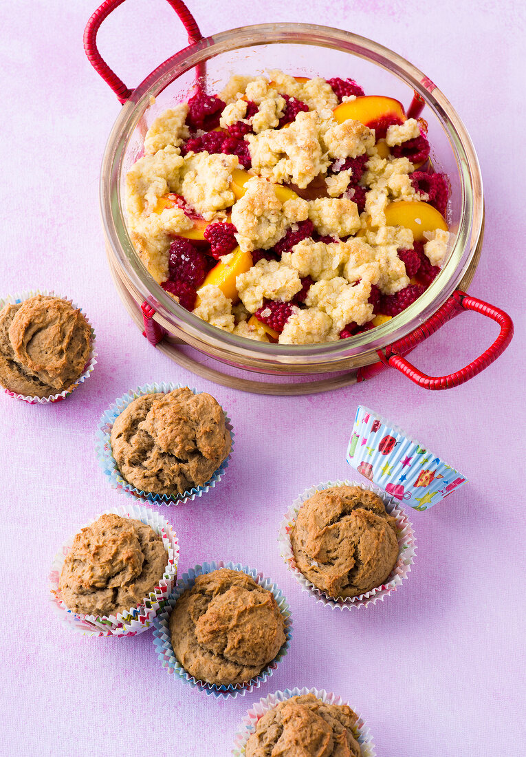Fruit crumble in glass bowl and muffins on purple background
