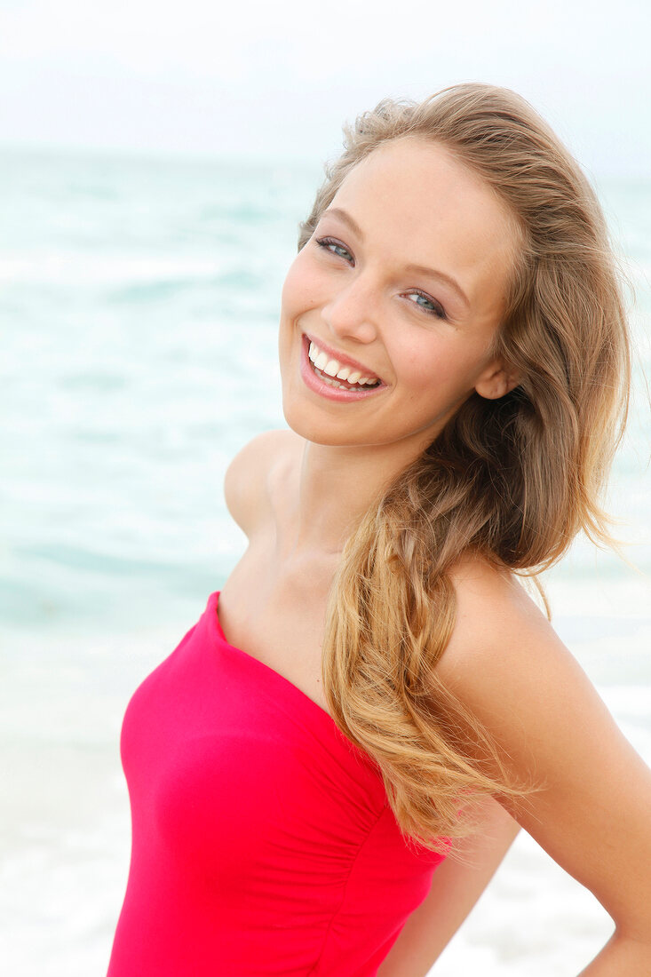 Portrait of beautiful blonde woman wearing red dress standing on beach, smiling