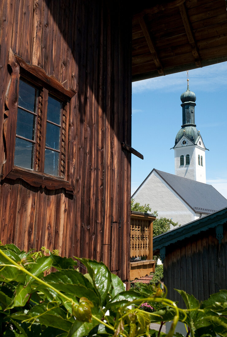 View of Stadtpfarrkirche Sankt Nikolaus through wooden house at Rimsting, Bavaria, Germany