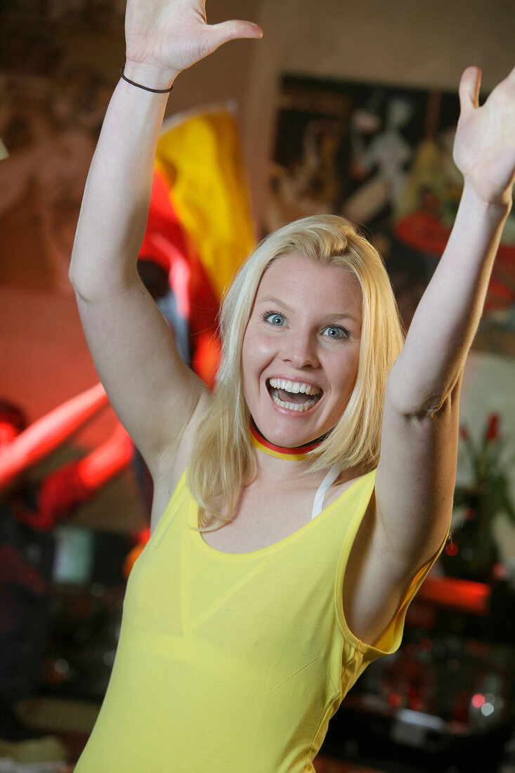 Blonde Frau bei Fußballübertragung, Top in gelb, hebt begeistert Hände