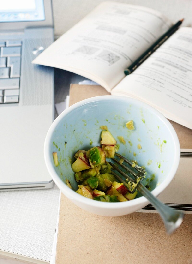Half eaten avocado salad with apples next to a laptop and book