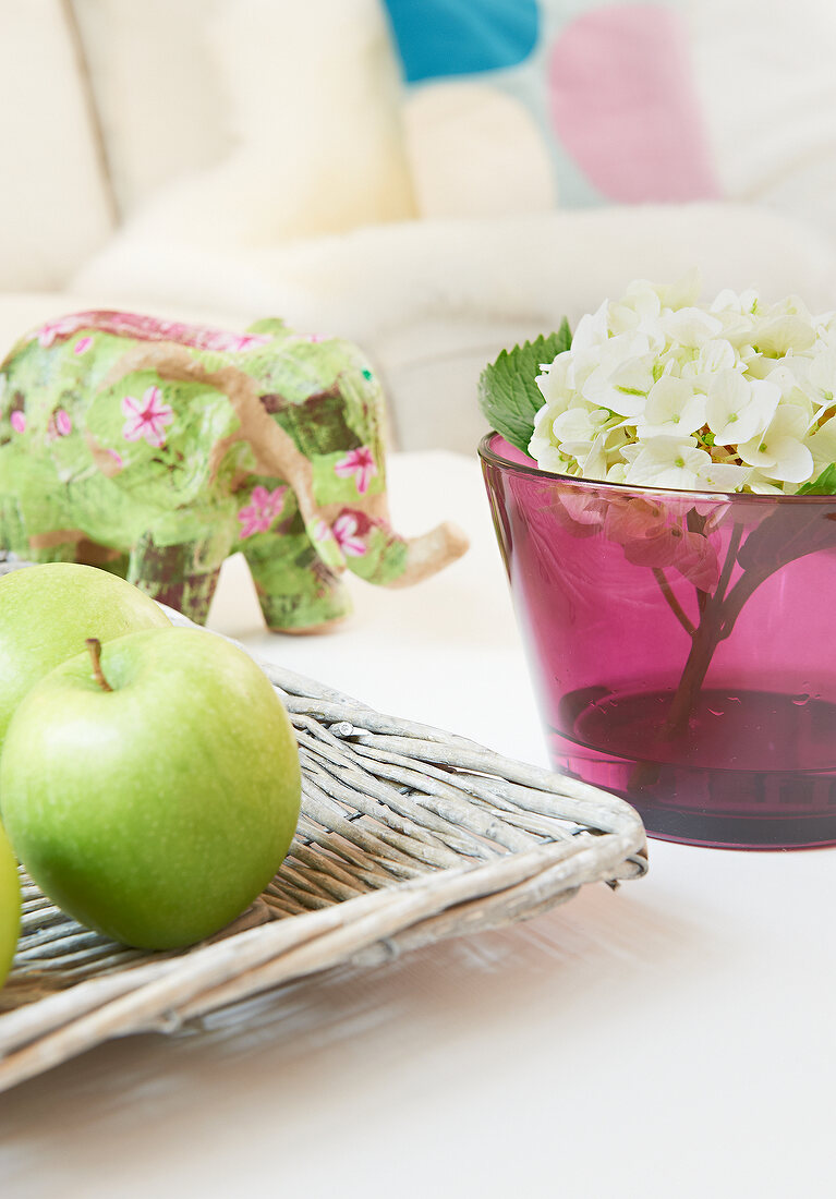 Close-up of wooden bowl with green apples, pink pot with flowers and decorative elephant