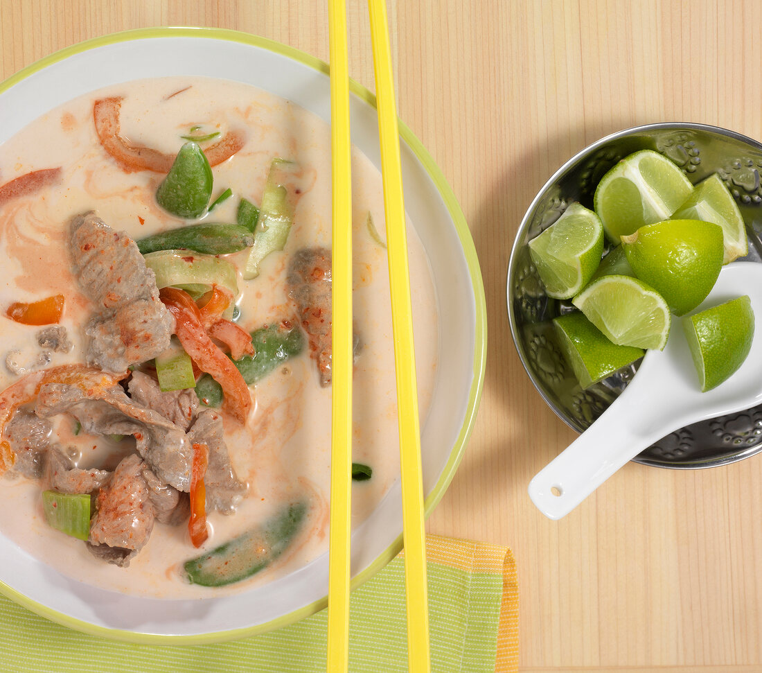 Coconut soup with beef in bowl with chopsticks on it and diced lemons in steel bowl